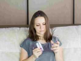 La foto ritrae una donna a letto che guarda una confezione di medicinali e un bicchiere d'acqua che tiene in mano. Il congedo non retribuito nei primi tre giorni di malattia potrebbe tornare per far fronte al debito pubblico sempre più in aumento.
