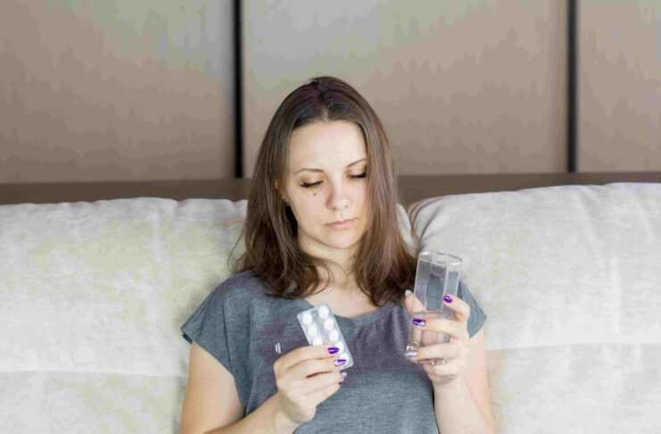 La foto ritrae una donna a letto che guarda una confezione di medicinali e un bicchiere d'acqua che tiene in mano. Il congedo non retribuito nei primi tre giorni di malattia potrebbe tornare per far fronte al debito pubblico sempre più in aumento.